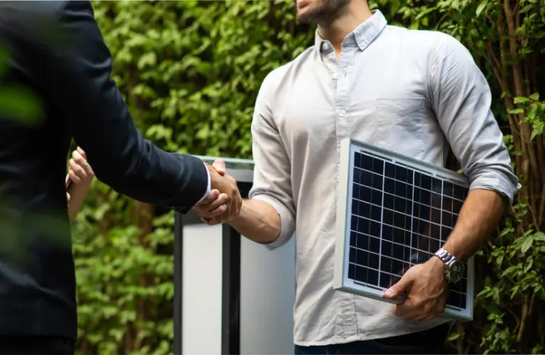 A professional handshake in progress between two individuals, one of whom is holding a small solar panel, symbolizing a green energy agreement or deal.