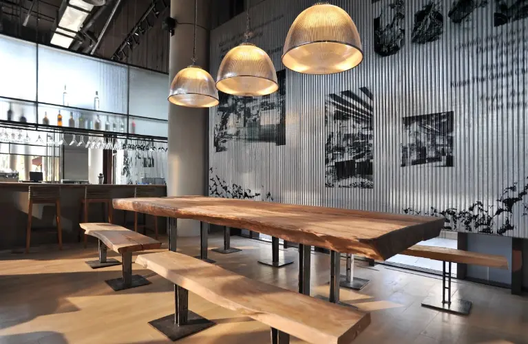 Modern commercial interior with a large wooden dining table, bench seating, industrial lighting, and corrugated metal wall paneling.