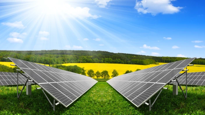 Dual rows of solar panels positioned on green grass with a blooming yellow field in the background, under a vivid blue sky with the sun radiating bright rays, illustrating eco-friendly energy production.
