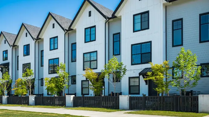 Exterior view of modern townhouses with white siding, black trim, and landscaped front yards featuring young trees and a black fence.